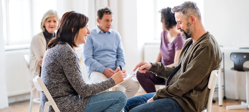 A senior counselor with clipboard talking to a man during group therapy.