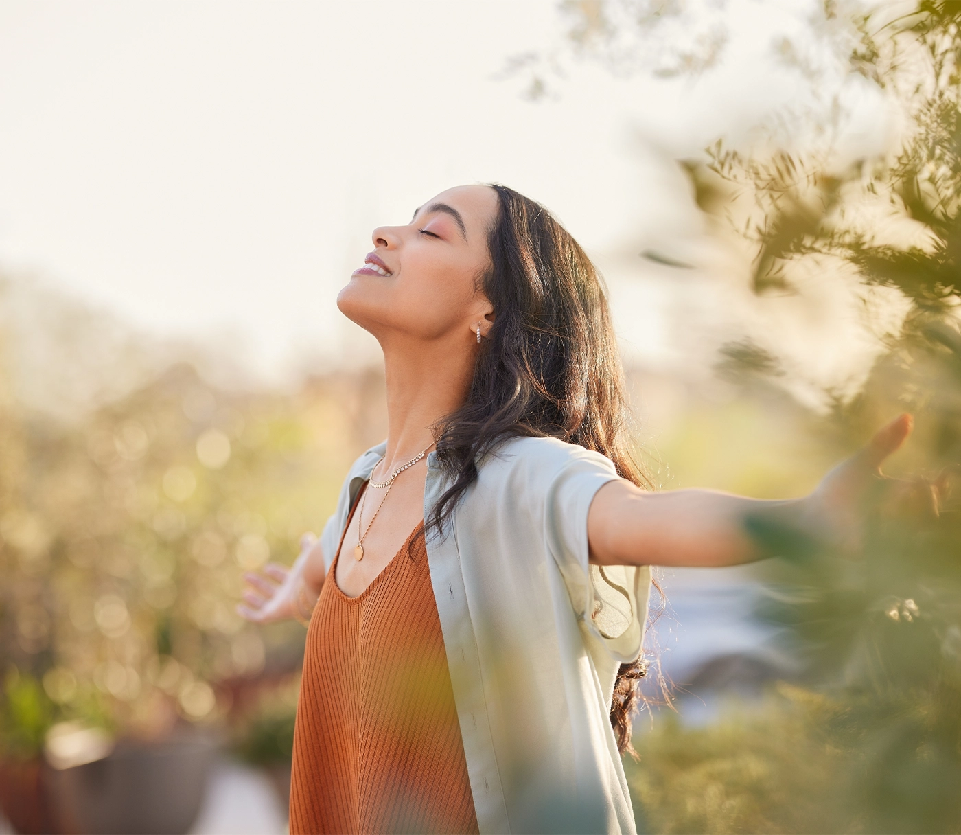 Young latin woman with arms outstretched breathing in fresh air during sunrise at the balcony. Girl enjoying nature while meditating during morning with open arms and closed eyes. Mindful woman relax.