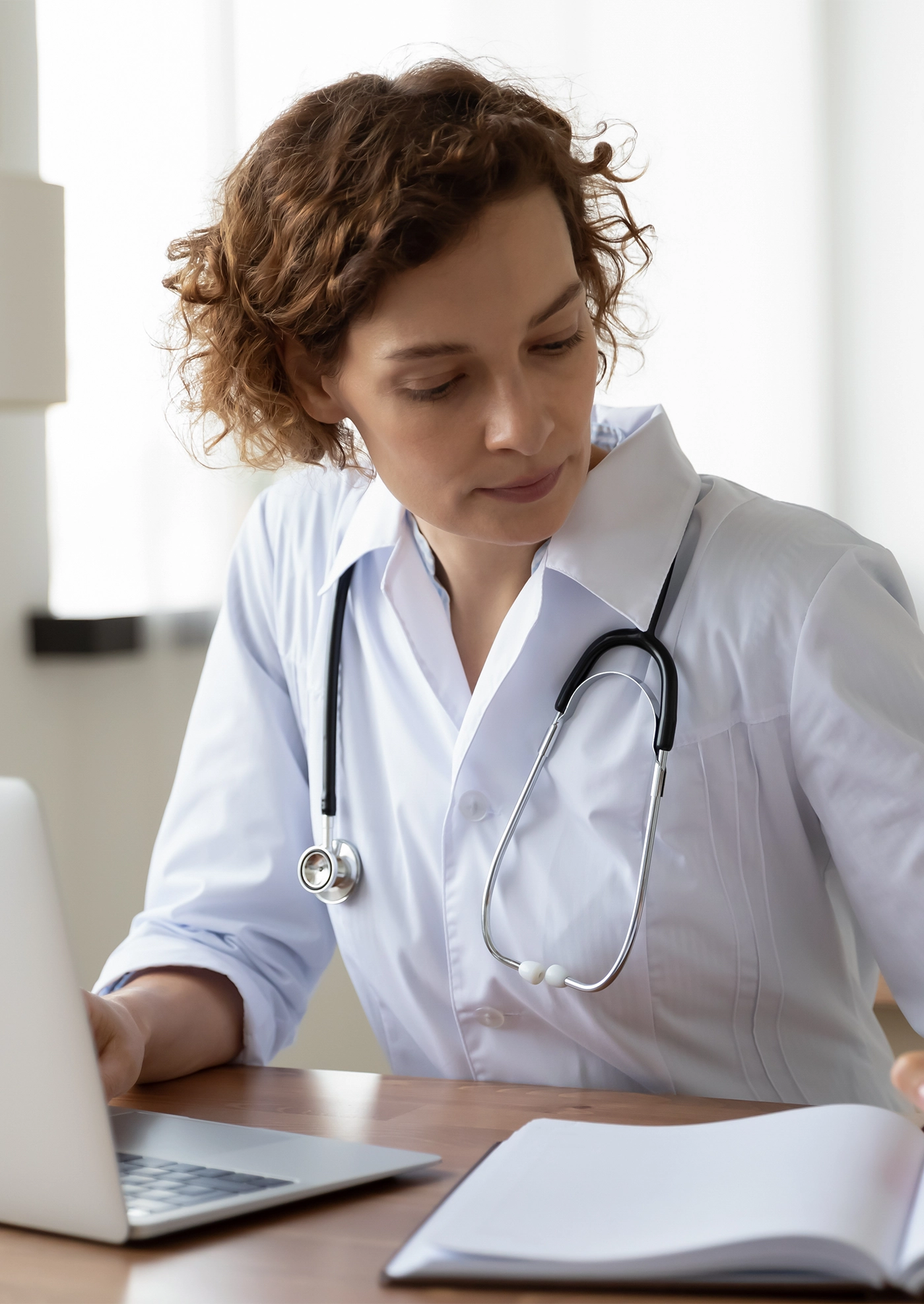 Serious female doctor using laptop and writing notes in medical journal sitting at desk. Young woman professional medic physician wearing white coat and stethoscope working on computer at workplace.