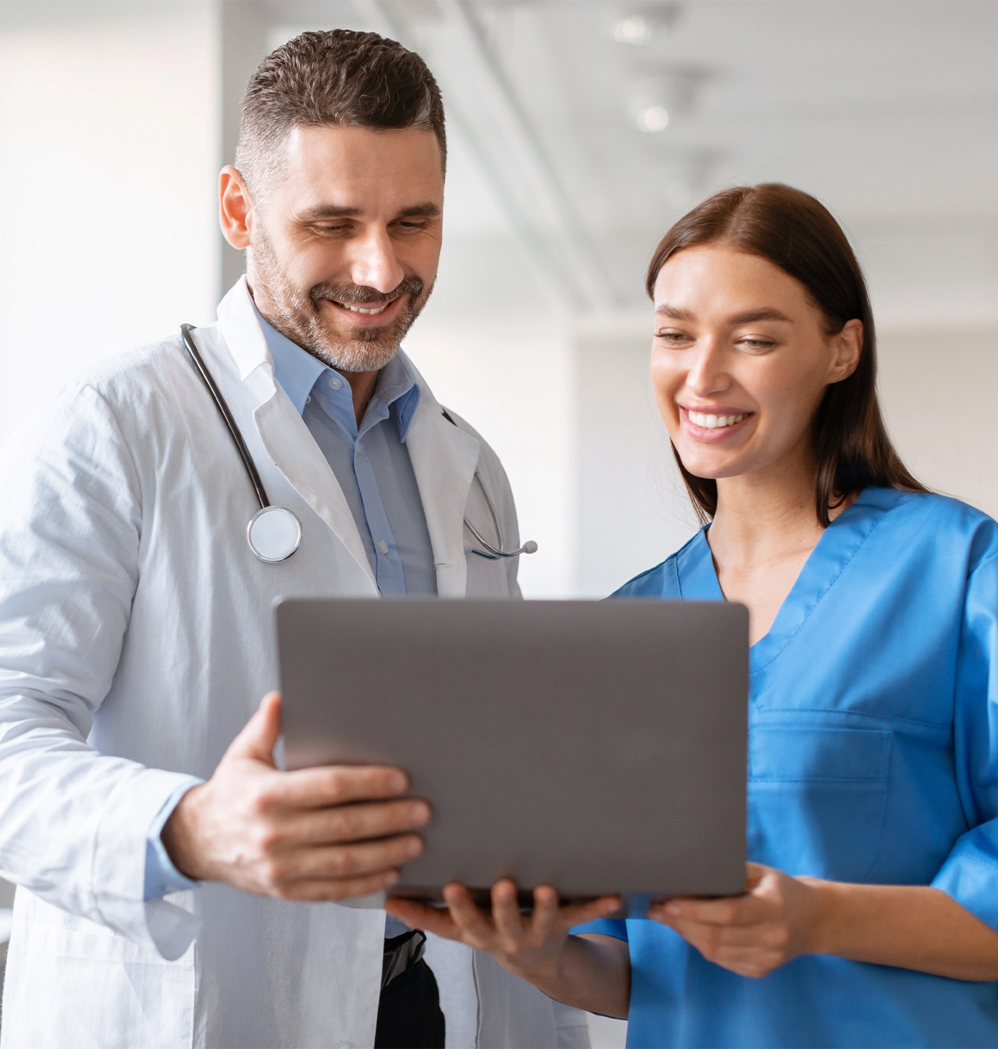 Happy male doctor and female nurse talking, working together, using laptop, discussing patient medical checkup results or diagnosis, wearing uniform holding electronic device
