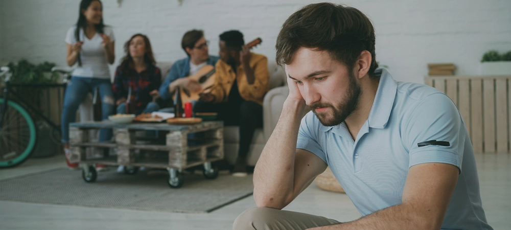 Young student guy feels upset and isolated while his friends celebrating party at home