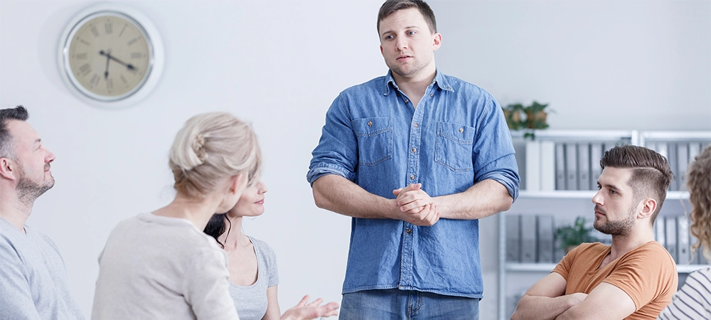 Guilty young man standing in an addiction therapy meeting and listening to psychologist
