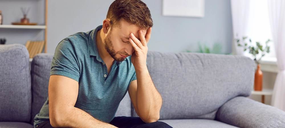 Portrait of upset sad depressed man sitting on sofa in living room at home alone suffering from headache. Young guy in bad thoughts feeling lonely indoors or having problems in relationships