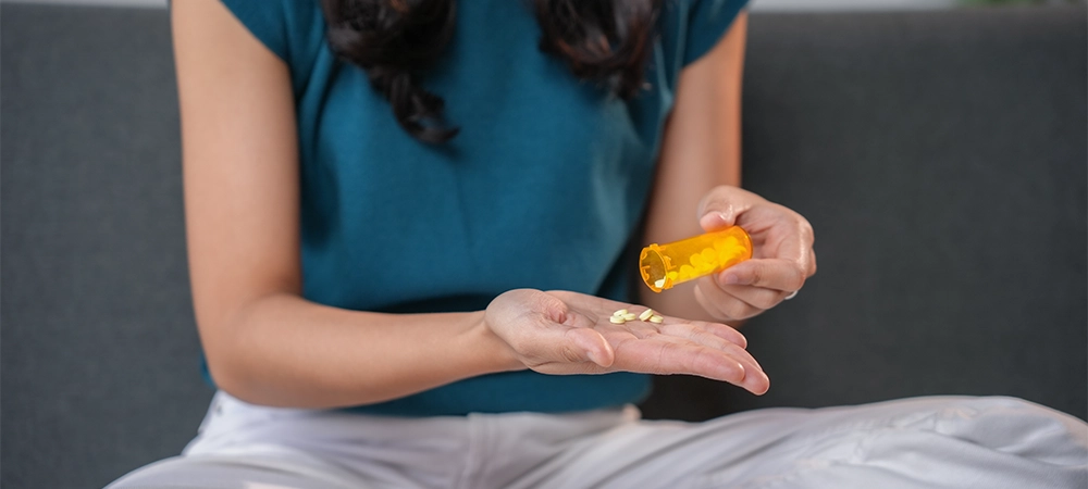 Young woman is pouring pills from a prescription bottle into her hand while sitting on a couch