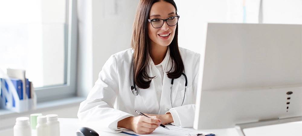 Cheerful woman medic professional giving online virtual consultation, making video call to patient and writing notes, sitting at desk and looking at computer monitor