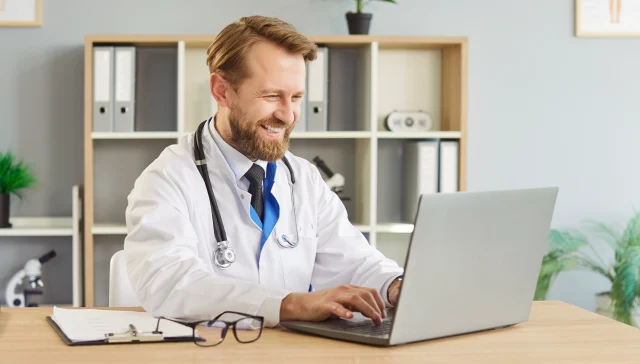 Doctor uses laptop for online telehealth consultation. Smiling male physician in modern clinic office reviews records and manages appointments remotely. Concept focus is remote medical care.