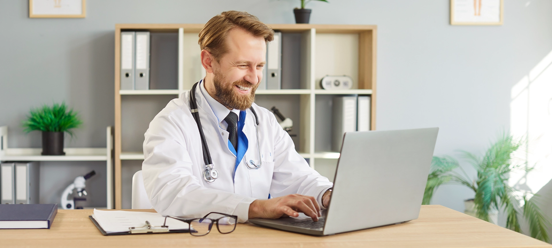 Doctor uses laptop for online telehealth consultation. Smiling male physician in modern clinic office reviews records and manages appointments remotely. Concept focus is remote medical care.