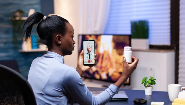Dark skinned woman showing pills bottle to doctor during video call late at night from living room. Black patient in a video call with medic discussing health problems of woman.
