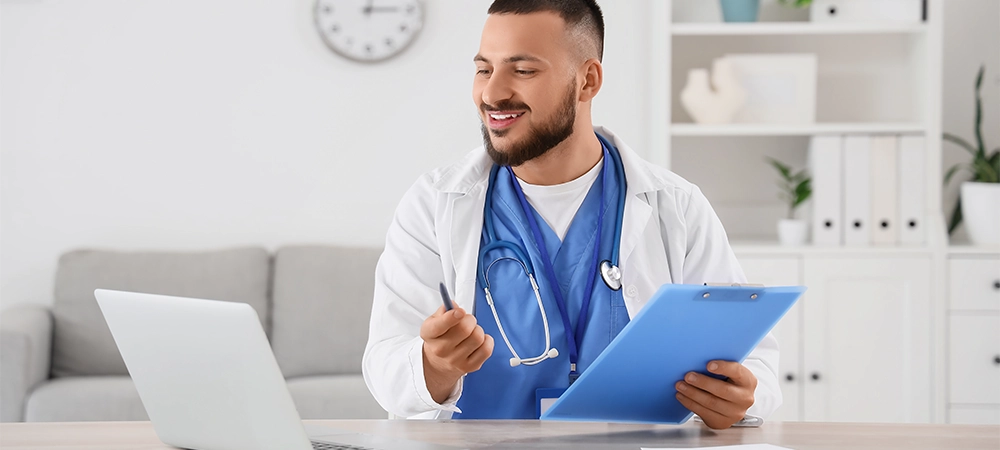 Male doctor with clipboard and laptop giving online consultation in clinic